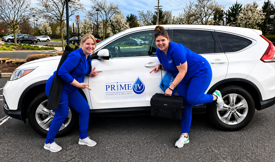 Two Prime IV nurses in blue scrubs pointing to the logo on the branded mobile-service car