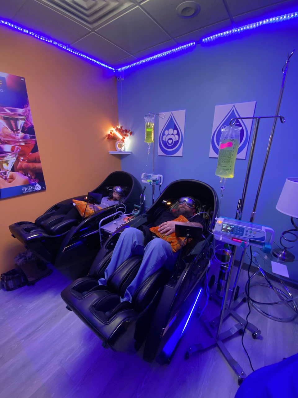 Two clients relaxing in massage chairs with light-therapy goggles under blue LED lighting in a private IV room