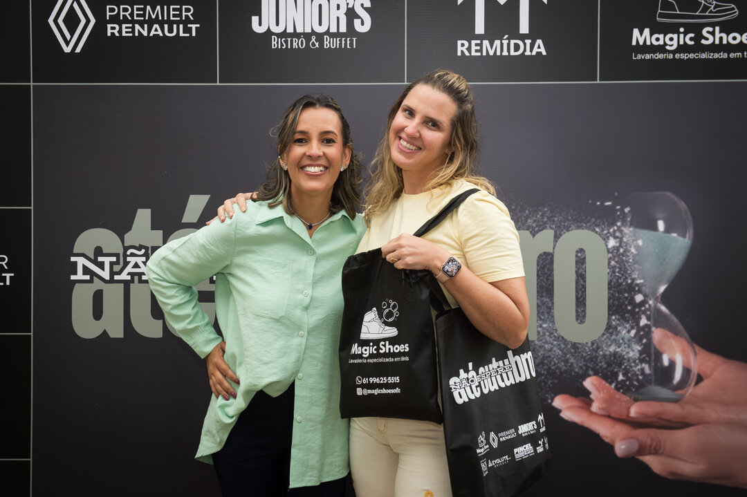 Duas mulheres com bolsas do evento em frente ao backdrop