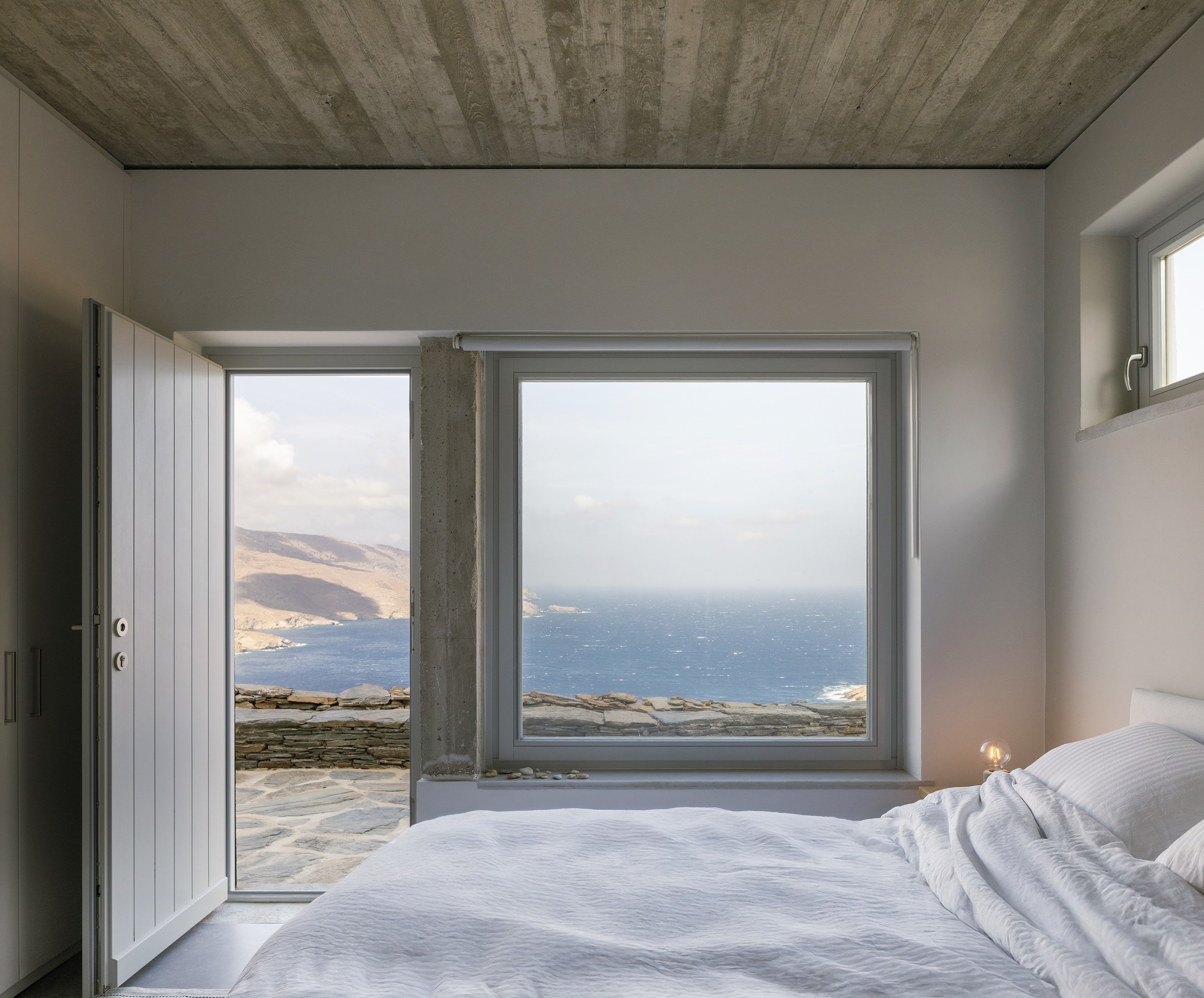 Bedroom with white linen bedding and a large window framing the Aegean Sea
