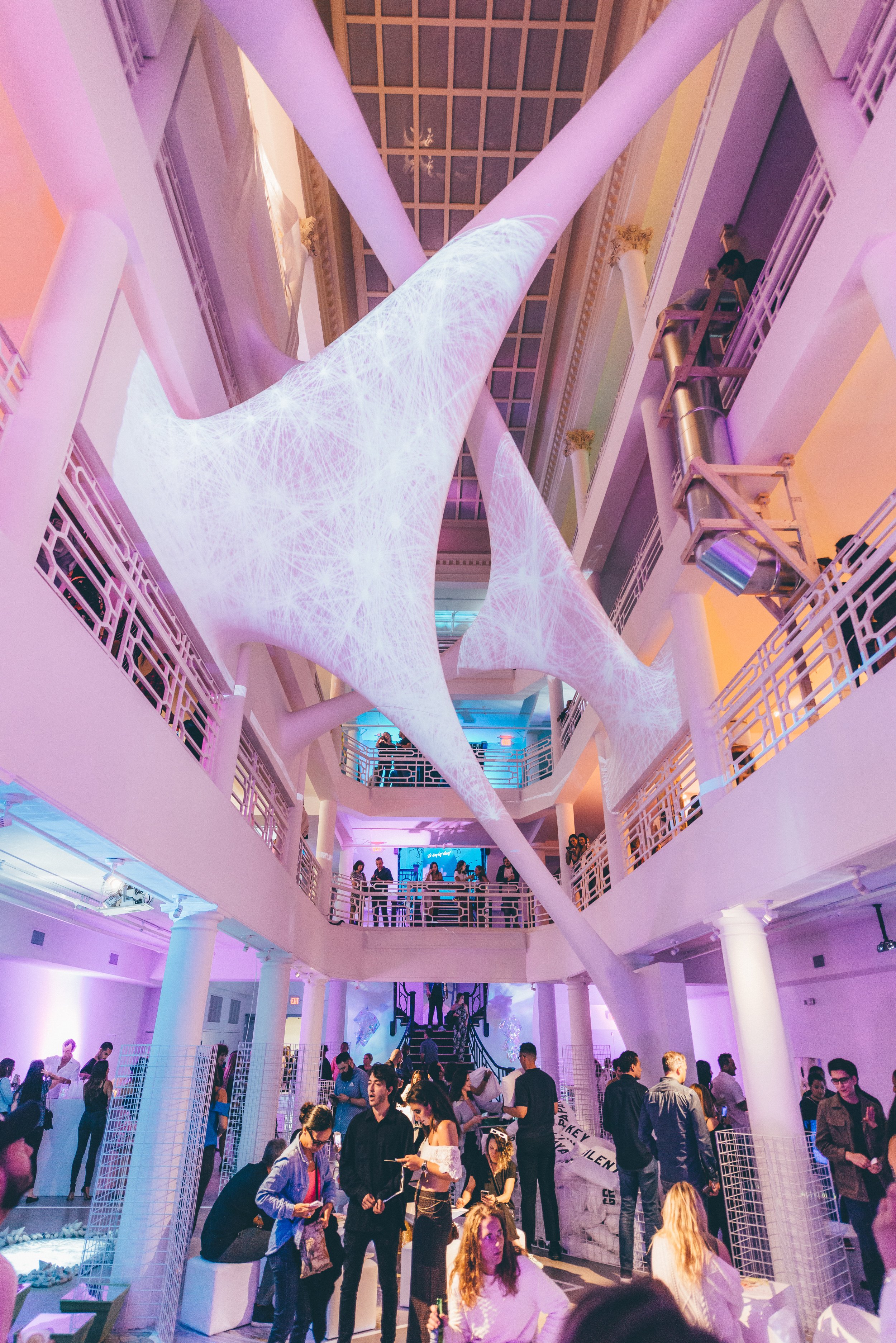 Large white sculptural installation suspended in a multi-story atrium with pink and purple lighting, crowds of visitors below