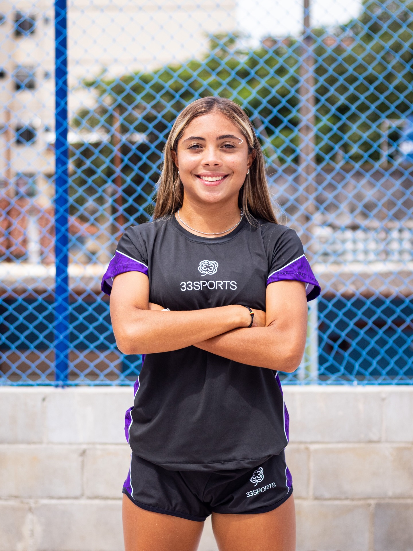 Atleta feminina de braços cruzados com uniforme preto e detalhes roxos 33Sports em quadra com tela azul