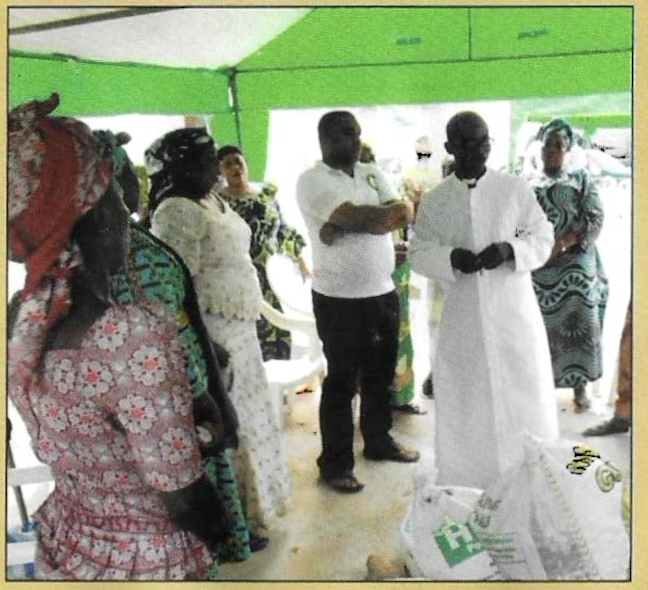 The Director and a priest with widows beside donated supplies under the canopy