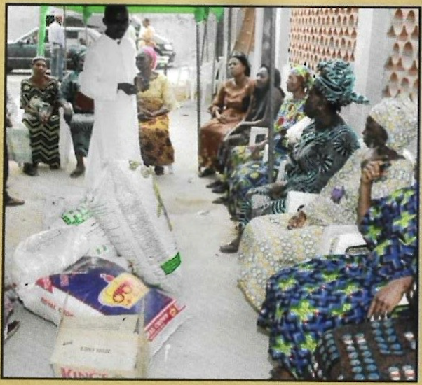 A priest addresses seated widows, food supplies laid out before them