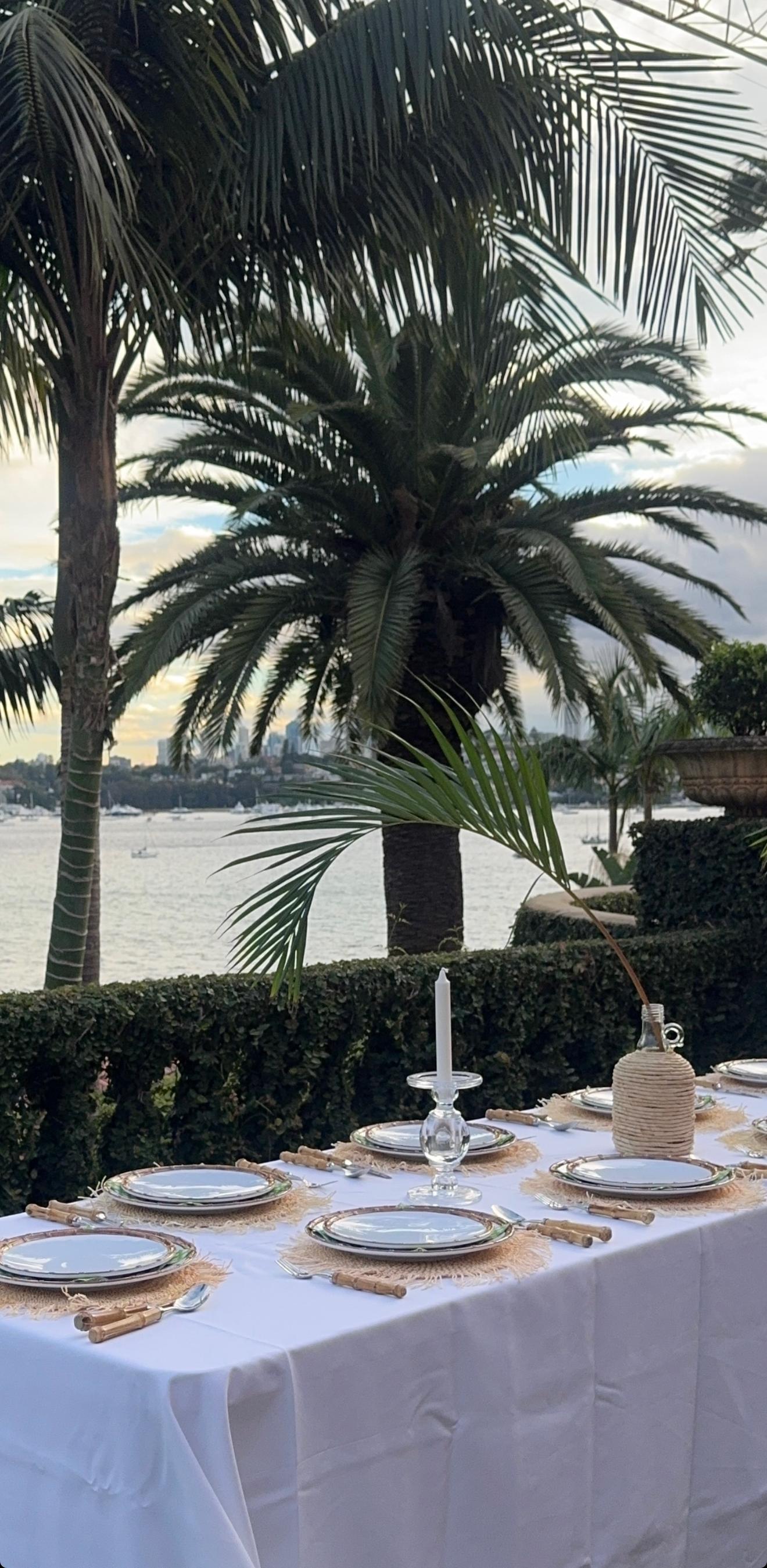 Elegant outdoor dining table with white linen, wicker placemats and crystal candlesticks overlooking Rose Bay harbour with palm trees