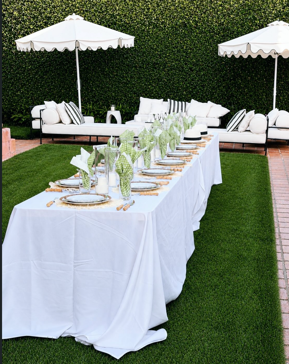 Outdoor event tablescape at Villa Biscaya, long white-draped table with green napkins and bamboo cutlery beneath scalloped white umbrellas beside white sofas against a manicured hedge