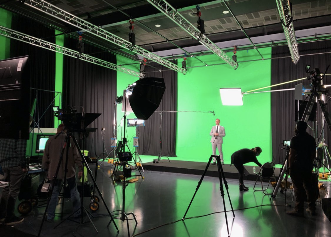 Film set with green screen, talent in a grey suit and a full camera and lighting crew