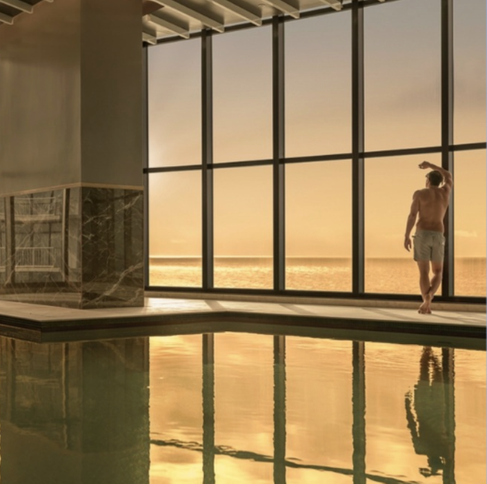 Editorial campaign location shot of a model by floor-to-ceiling windows at golden hour beside an infinity pool