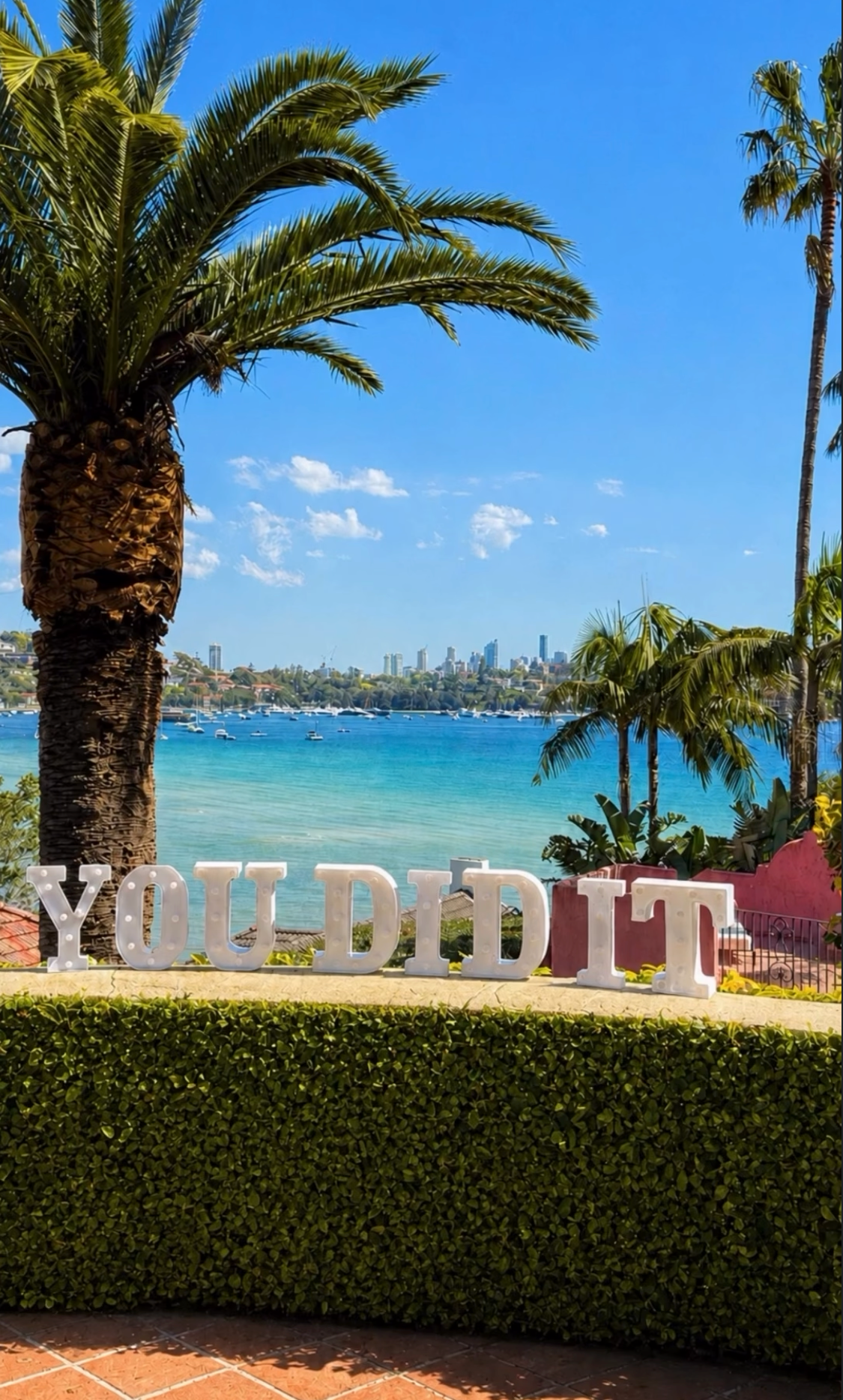 Large illuminated YOU DID IT marquee letter sign on the Villa Biscaya lawn hedge with Sydney Harbour, palm trees and blue sky behind