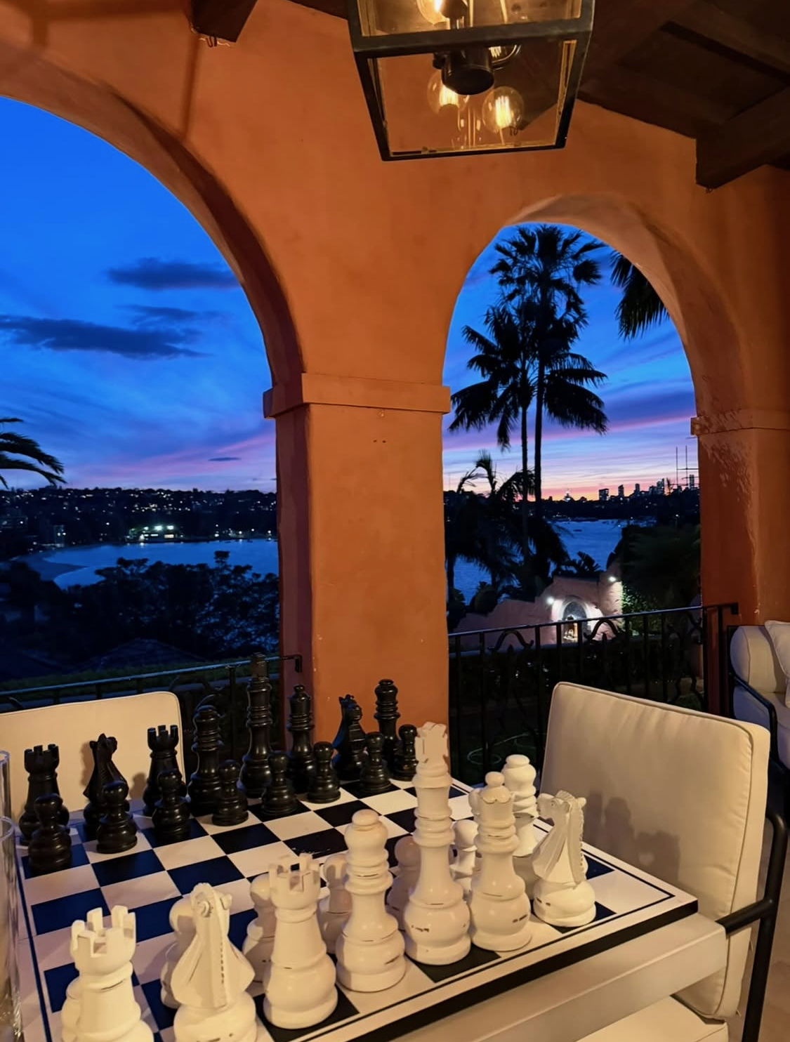 Large chess set on loggia table at deep blue and pink dusk sunset, terracotta arch with hanging lantern, harbour and palm silhouettes beyond