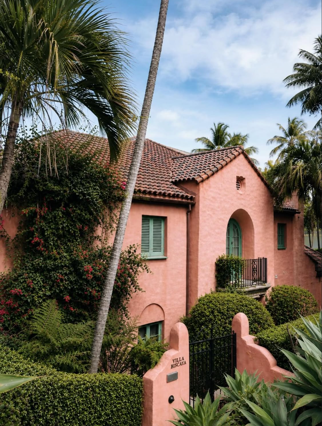 Villa Biscaya entrance gate with pink Spanish Mission facade, terracotta tile roof, green shutters, bougainvillea and lush tropical garden with palm trees