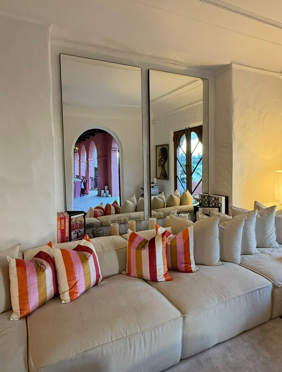 Villa Biscaya interior living room with cream sectional sofa, colourful striped cushions, two tall floor mirrors reflecting the pink arched loggia and coffee table books