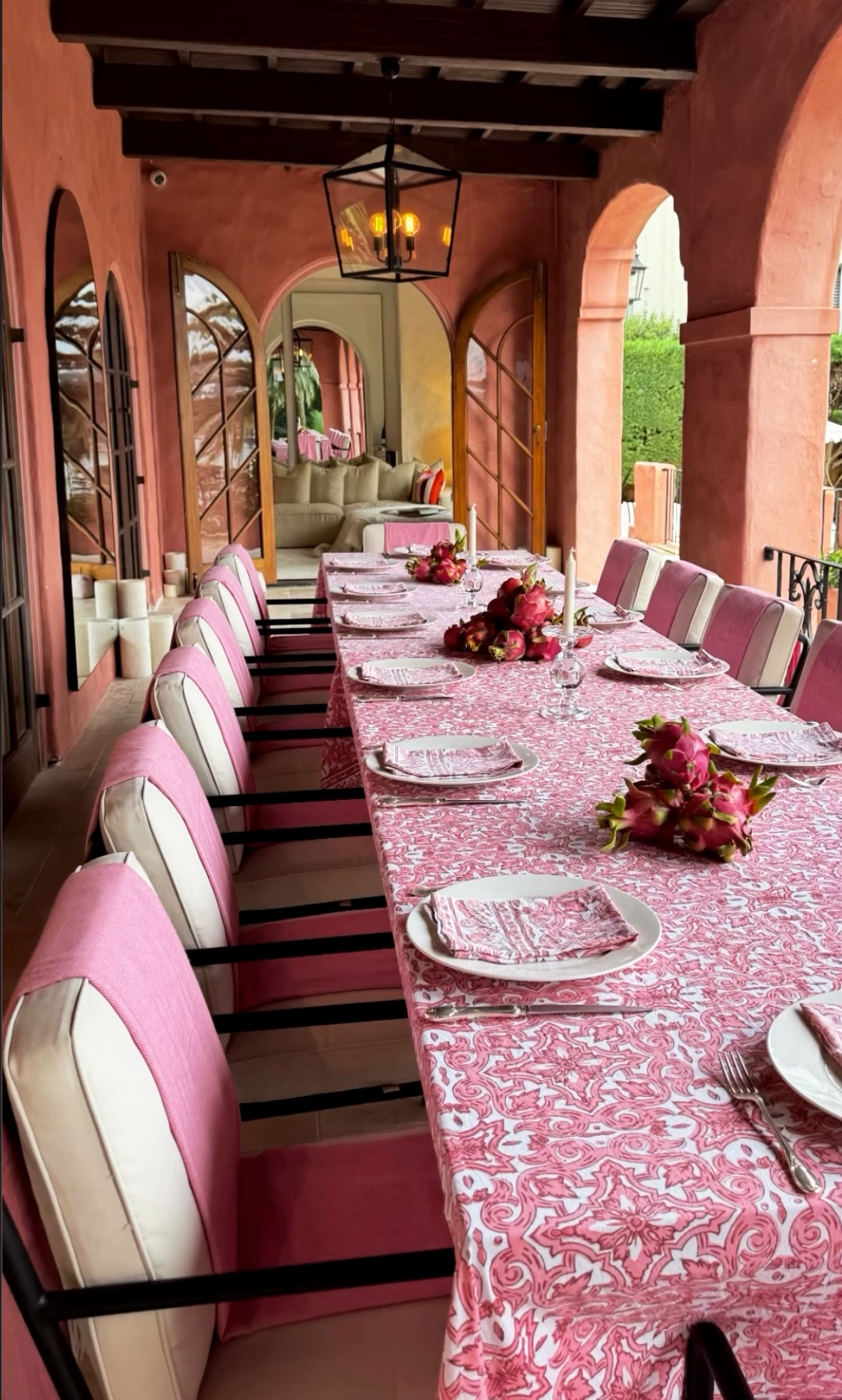 Loggia long dining table set with pink damask tablecloth, pink velvet chairs, dragon fruit centrepieces and hanging lantern, arched doorways opening to the living room