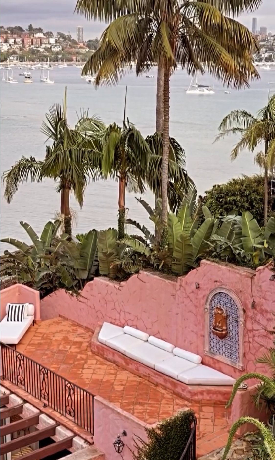 Pink rooftop terrace with white daybeds and striped pillow, mosaic fountain niche in the pink wall, lush tropical garden and Sydney Harbour with sailboats behind