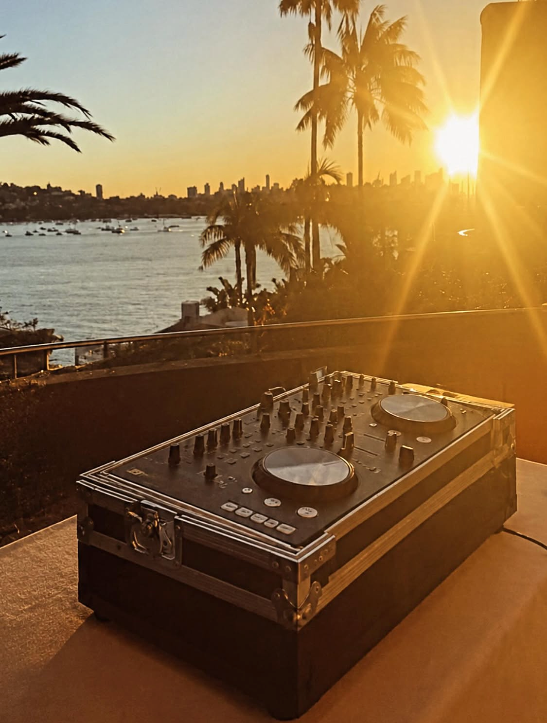DJ decks on Villa Biscaya terrace at golden harbour sunset with palm tree silhouettes, Sydney skyline and dramatic sun flare