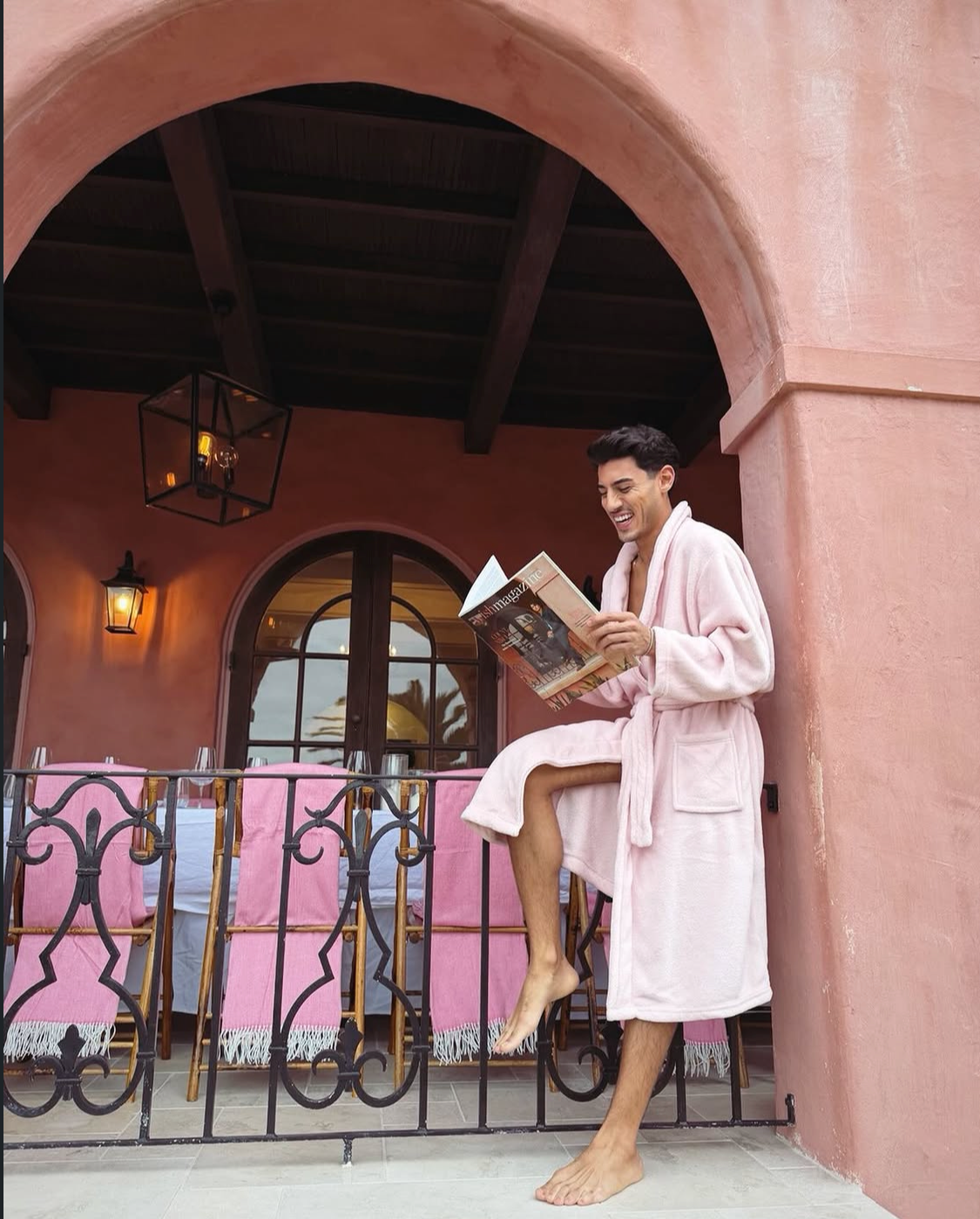 Man in a pink bathrobe seated on the iron railing reading Wish Magazine, pink bamboo chairs and arched loggia with lantern behind