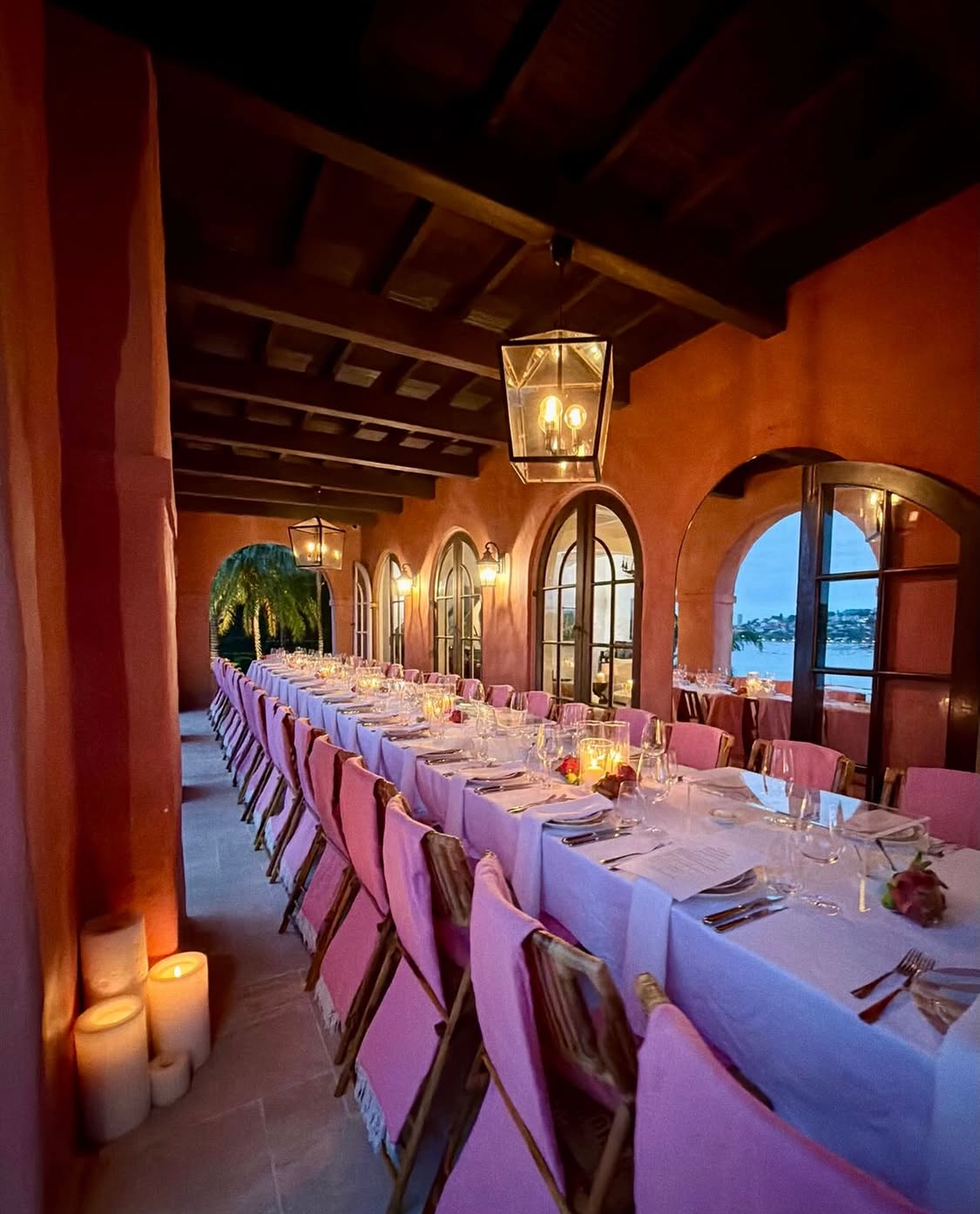 Grand candlelit loggia dinner at dusk with long white-linen table, pink bamboo chairs, pillar candles on the floor, hanging lanterns and arched windows framing the harbour blue glow and palm trees