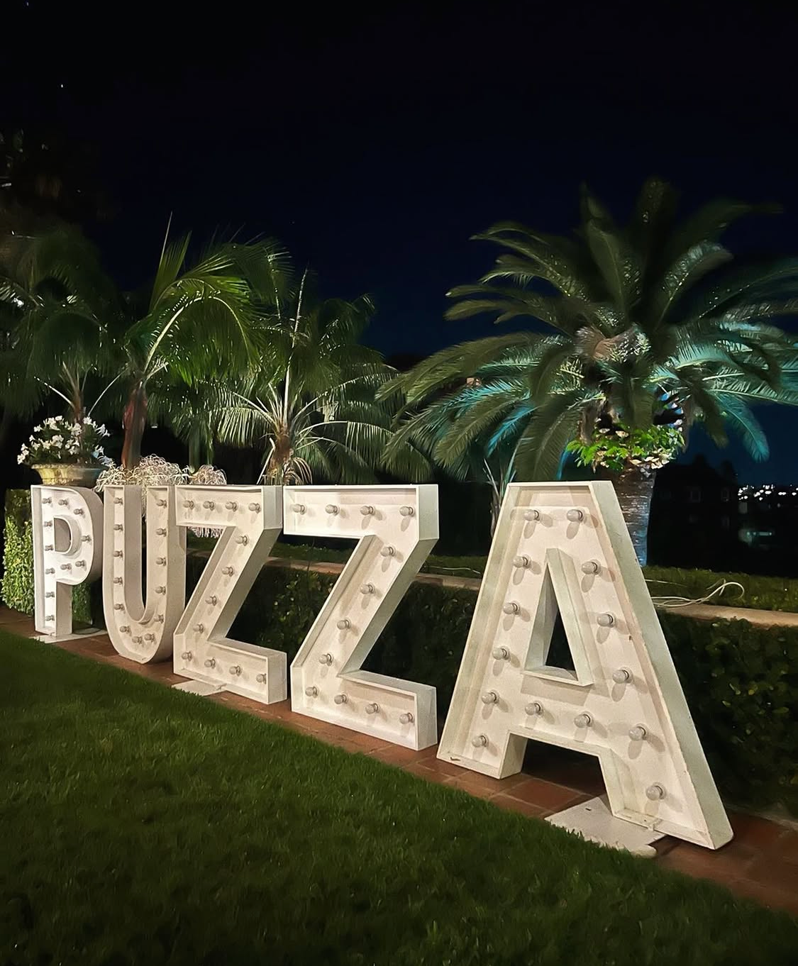 PUZZA large illuminated marquee letter sign on the Villa Biscaya lawn at night, tropical palms lit from below against a deep blue sky