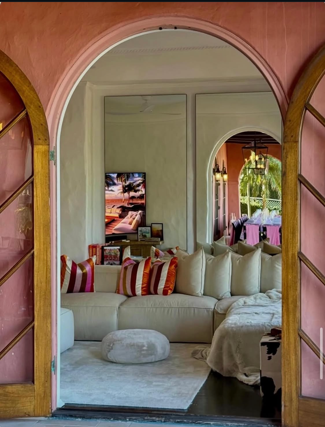Interior living room viewed through open golden arched French doors, cream sectional sofa with bold striped cushions, floor mirrors, TV showing sunset artwork and velvet pouf on a white rug