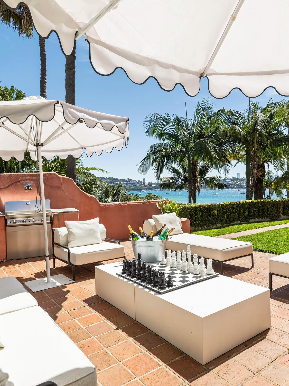 Poolside terrace under scalloped white umbrellas with chess set and champagne bucket on white cube table, white daybeds, pink estate wall and Sydney Harbour with palm trees