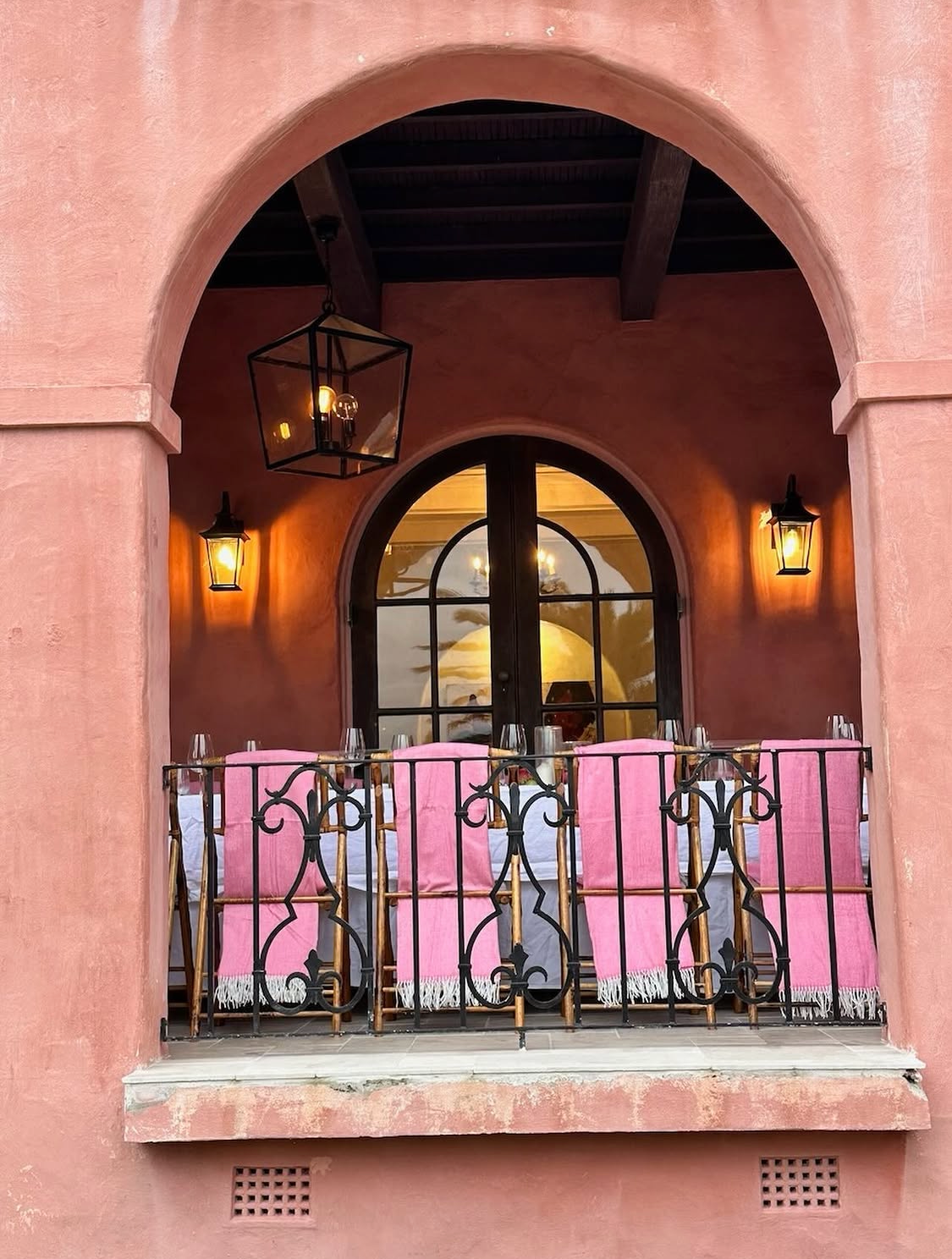 Villa Biscaya loggia arch exterior at dusk, pink bamboo chairs with pink throws and crystal glassware visible through the ornate iron railing, hanging lantern and wall sconces glowing warmly