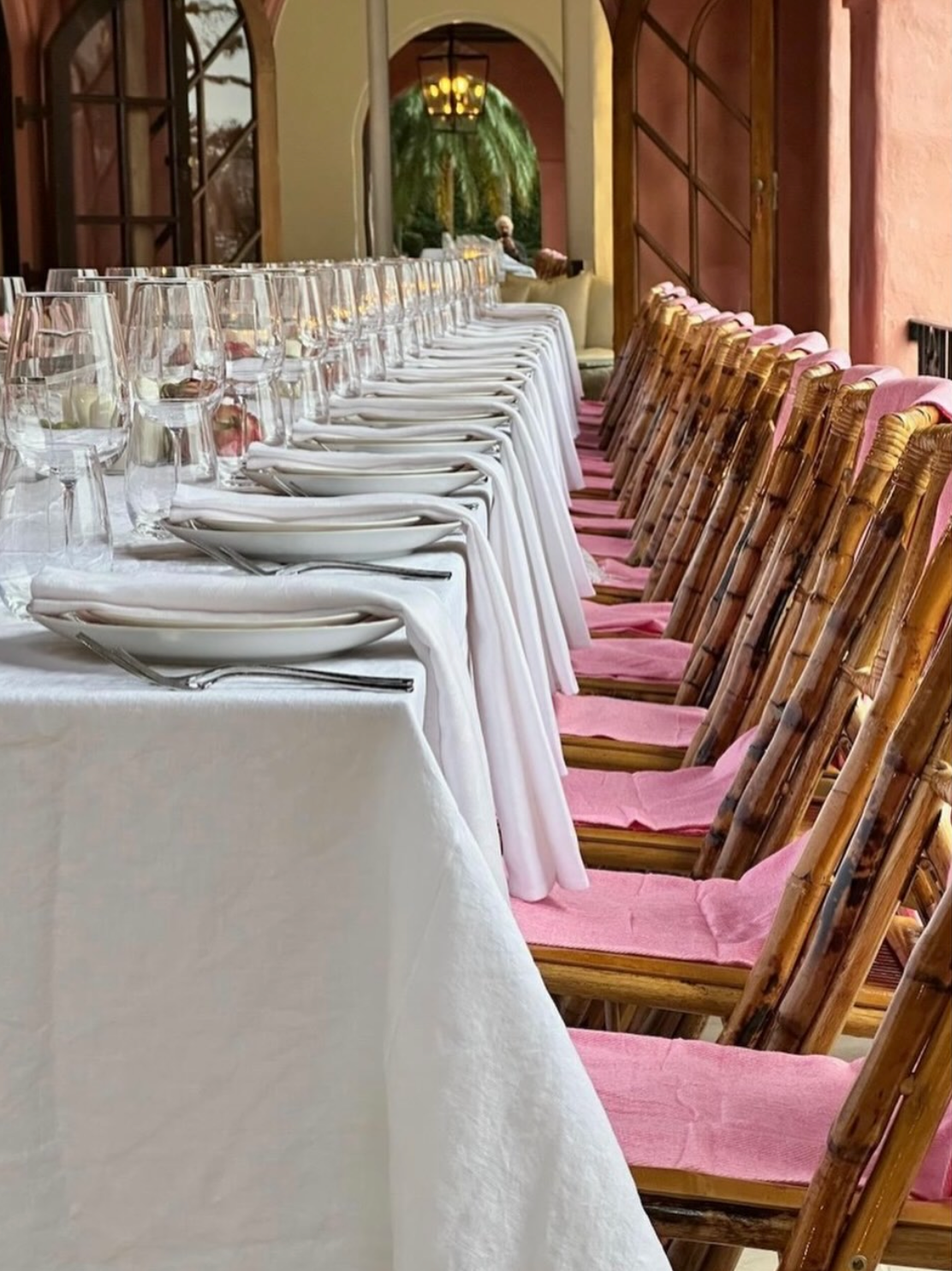 Close-up of long loggia dining table with crisp white linen, pink bamboo chairs with fringe throws and rows of crystal wine glasses, terracotta archways receding into the distance