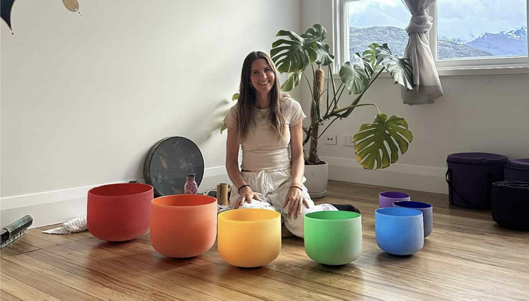 Julie Faulkner kneeling beside a rainbow of crystal singing bowls in a bright, plant-filled room
