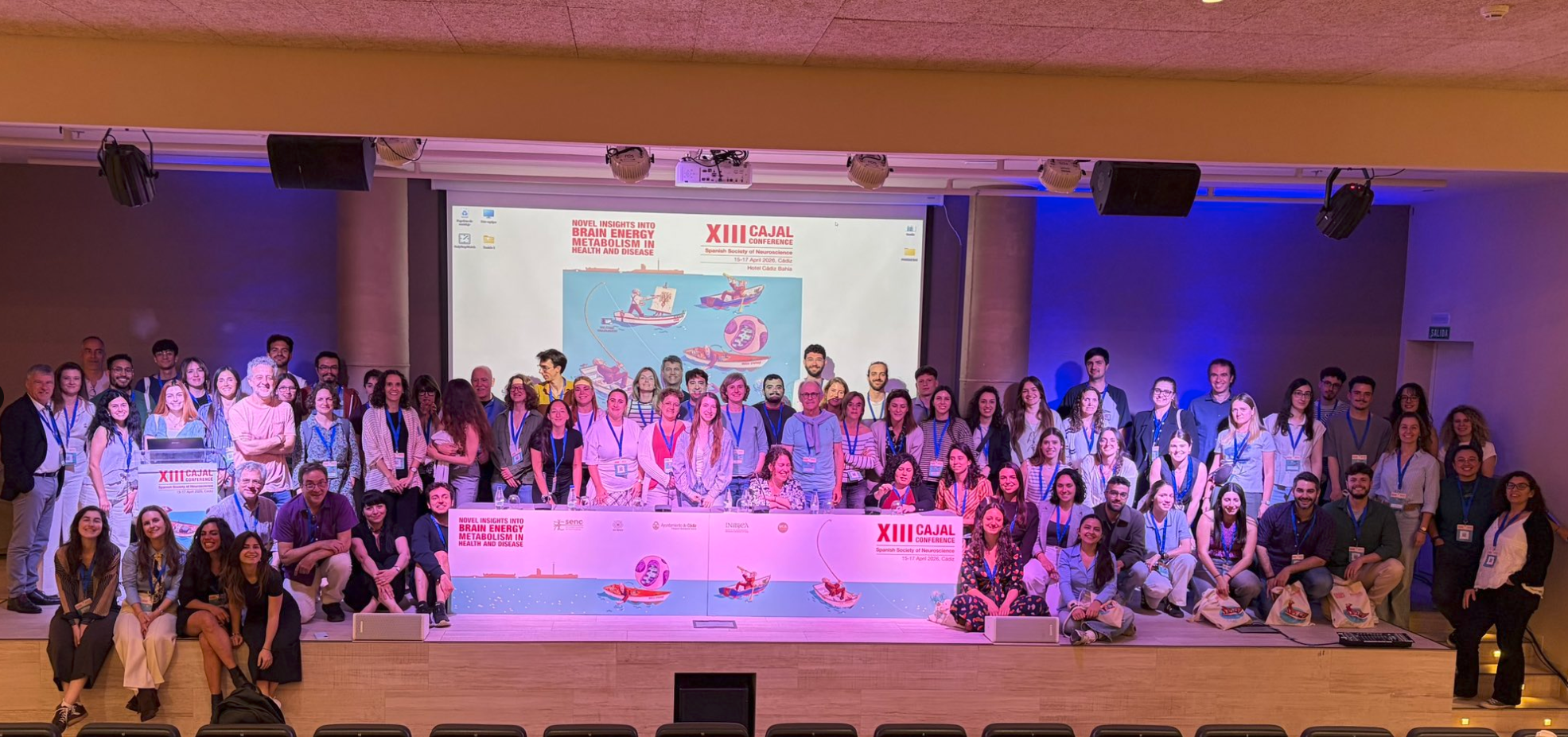 Group photo of all XIII Cajal Conference attendees on stage with purple lighting and conference banner