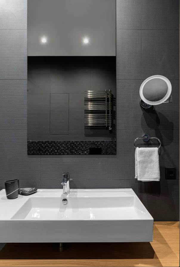 Dark gray mosaic tile bathroom with large rectangular white floating sink, recessed mirror niche with black mosaic accent, chrome towel radiator, round illuminated wall mirror, wood floor