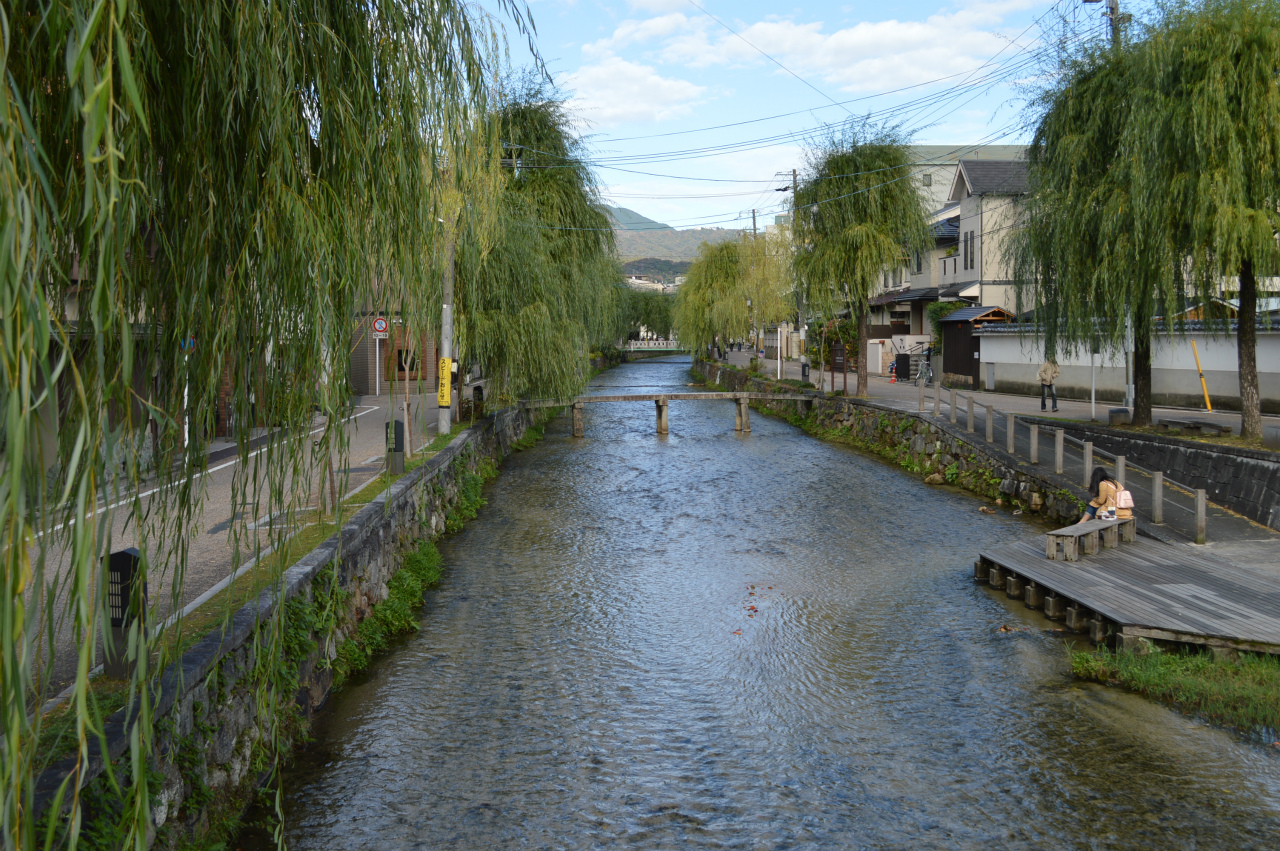 Shirakawa Canal lined with weeping willow trees, stone embankments, wooden deck platform, and Higashiyama mountains in background