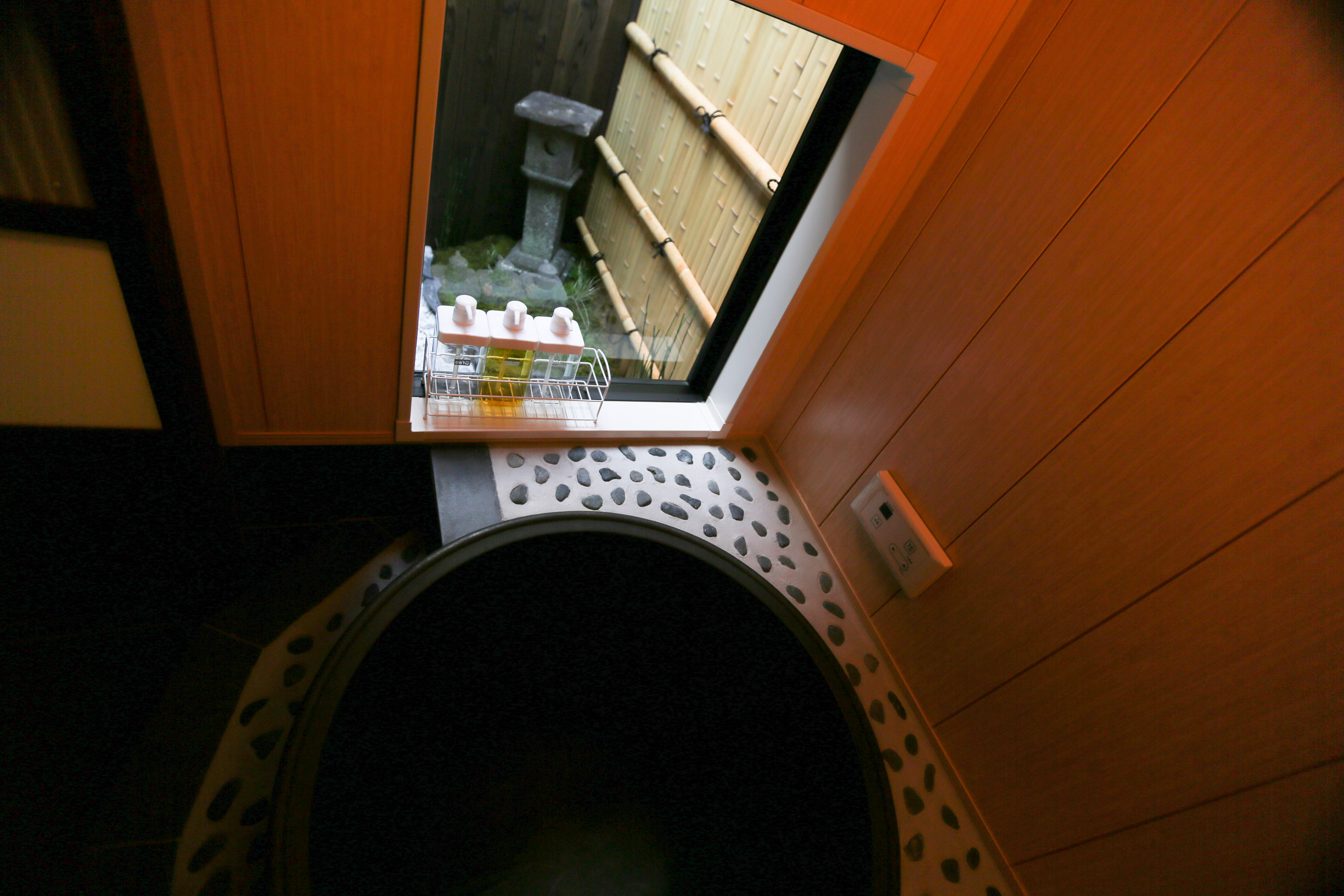 Bathroom with deep black goemon soaking tub, hinoki wood paneling, river pebble accents, and garden view window with bamboo fence