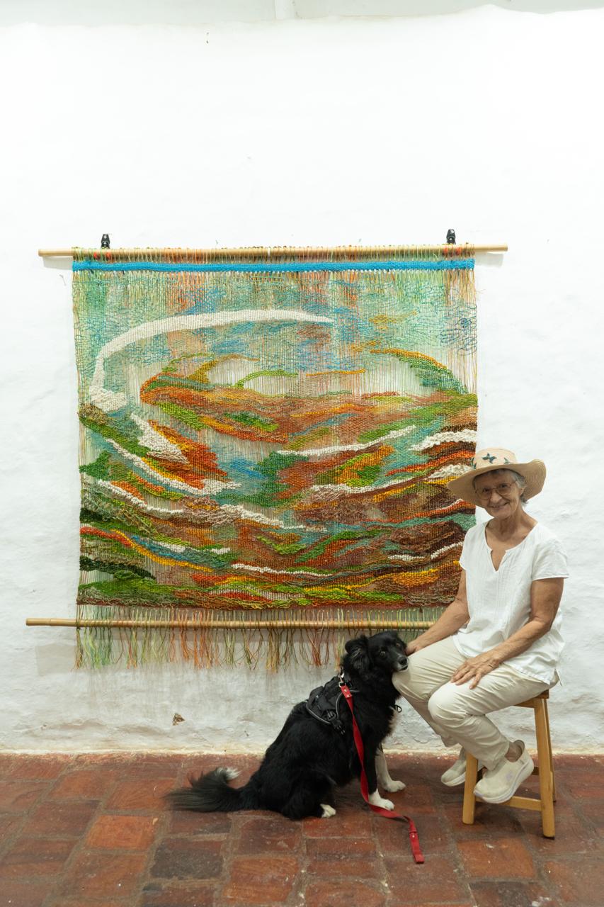 Silvia Medina con su perro Dalí junto a un gran tapiz tejido con paisaje