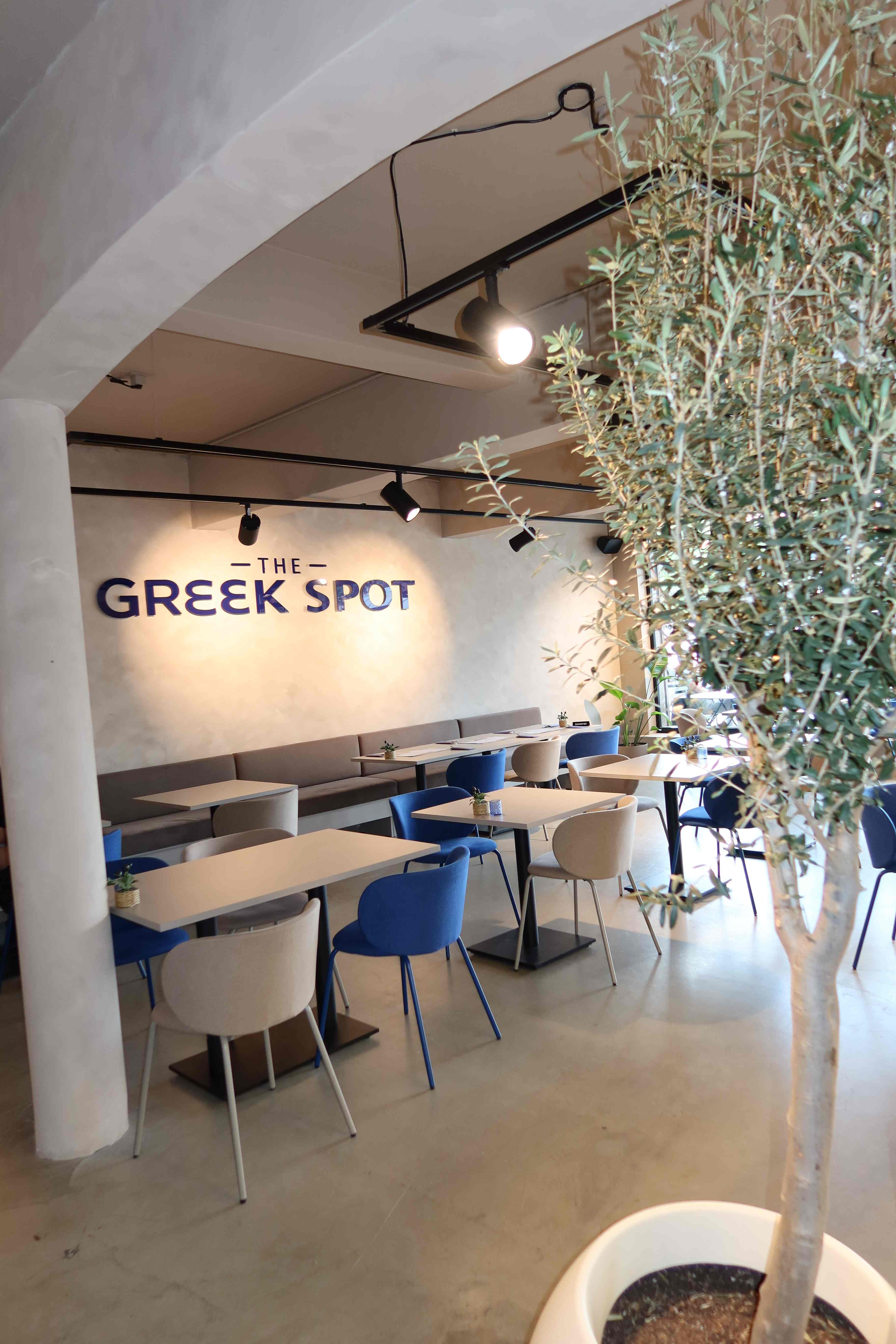 Dining area with blue and beige chairs, olive tree, and The Greek Spot logo on the wall