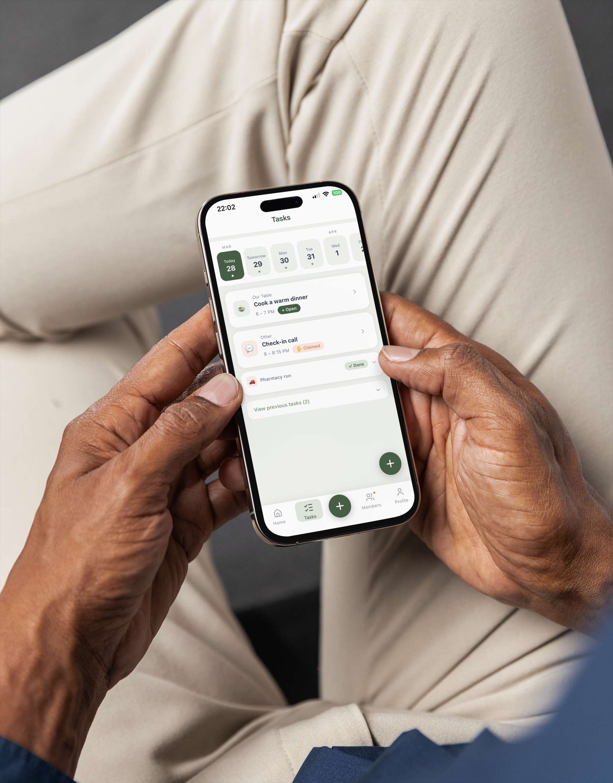 Hands holding a phone showing the VillageLink Tasks interface with scheduled items like 'Cook a warm dinner', 'Check-in call', and 'Pharmacy run'