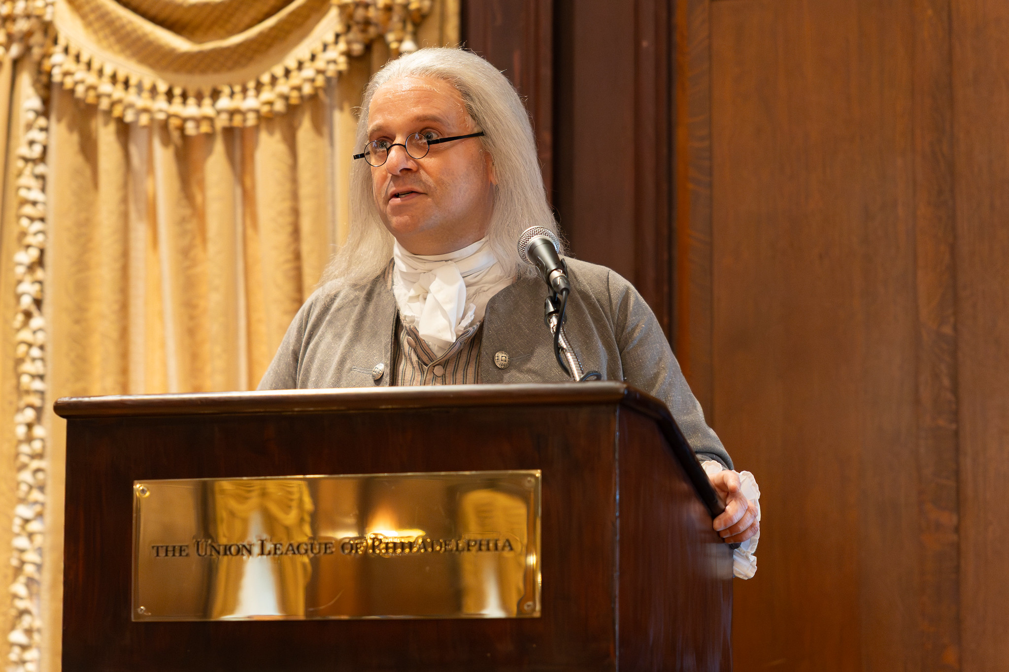 A teacher in colonial Benjamin Franklin costume delivering a living-history presentation at a podium