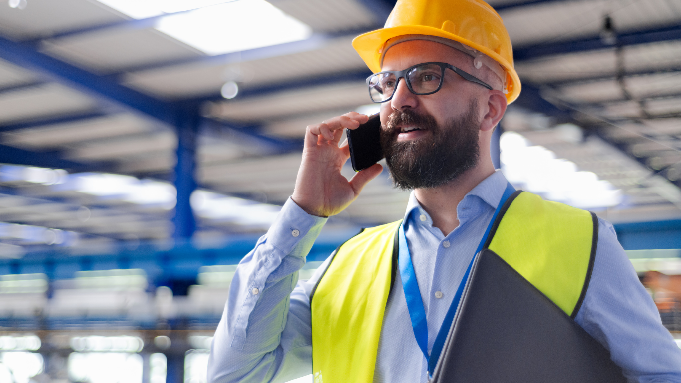Industrial safety professional in hard hat and safety vest on phone in warehouse