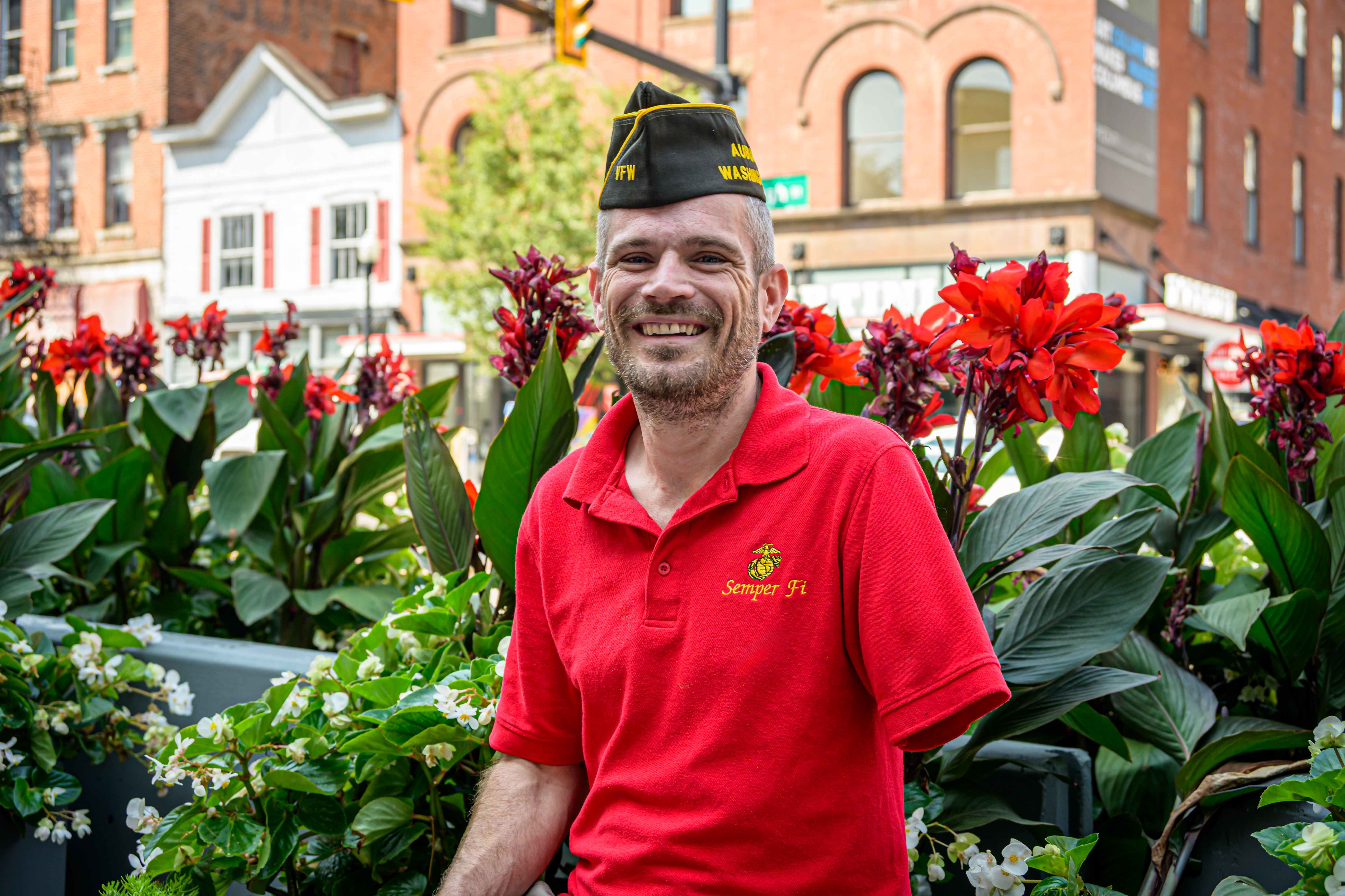 Marine Corps veteran in red Semper Fi polo and VFW garrison cap smiling in urban setting