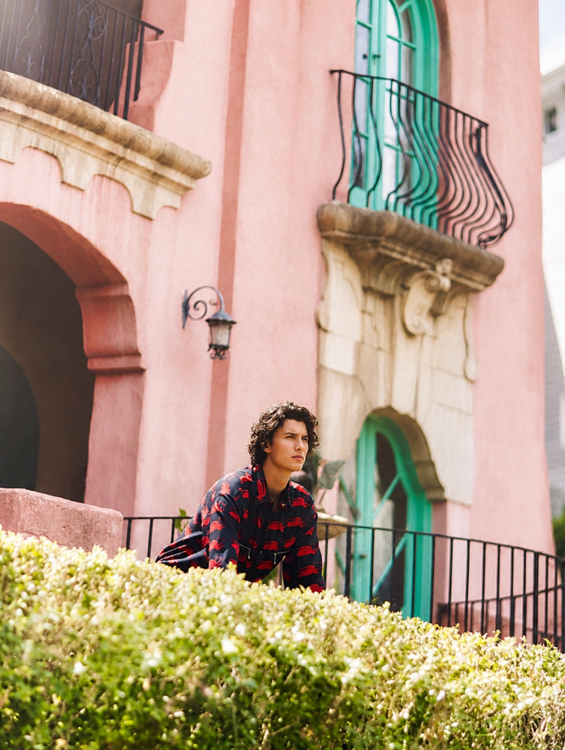 Count Nikolai in a dark navy and red printed shirt leaning over the black iron railing in front of Villa Biscaya's pink facade, with the green arched doorway, ornate sandstone balcony corbel and trimmed hedges in the foreground