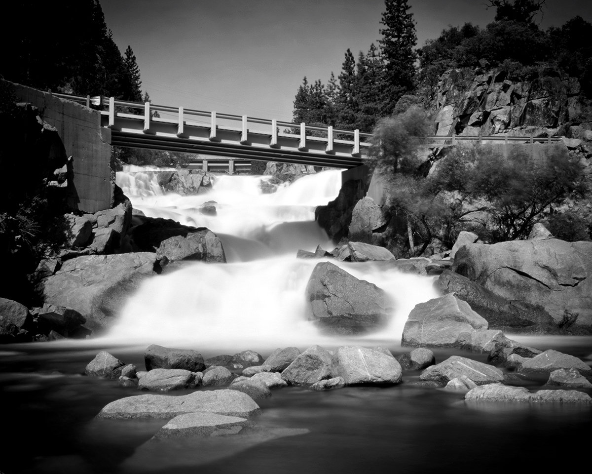 Cascading waterfall beneath white bridge with rocks, forest backdrop, and dreamlike long exposure water flow