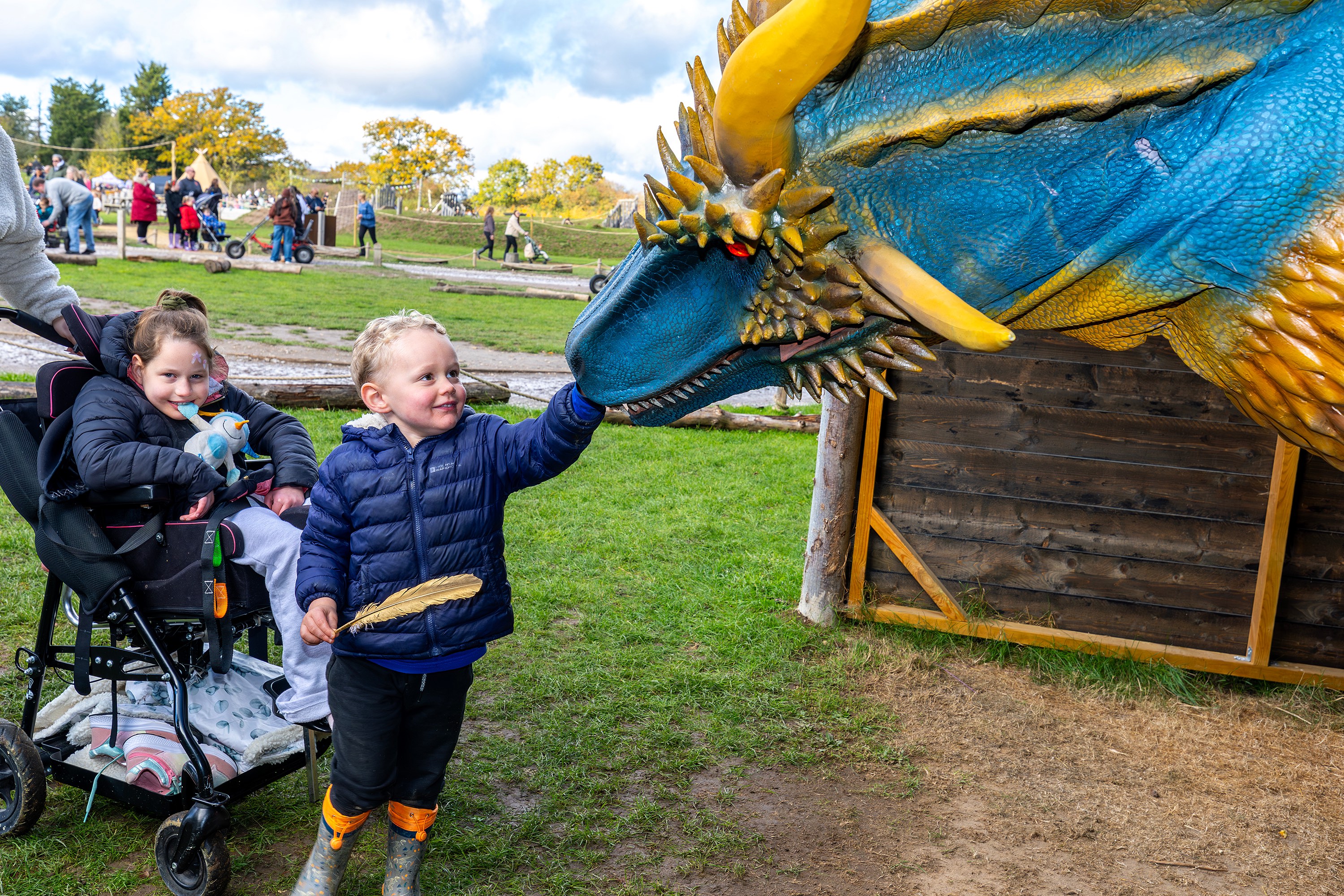Child in wheelchair and sibling reaching out to touch the blue dragon at Wyvernwood SEND Fest