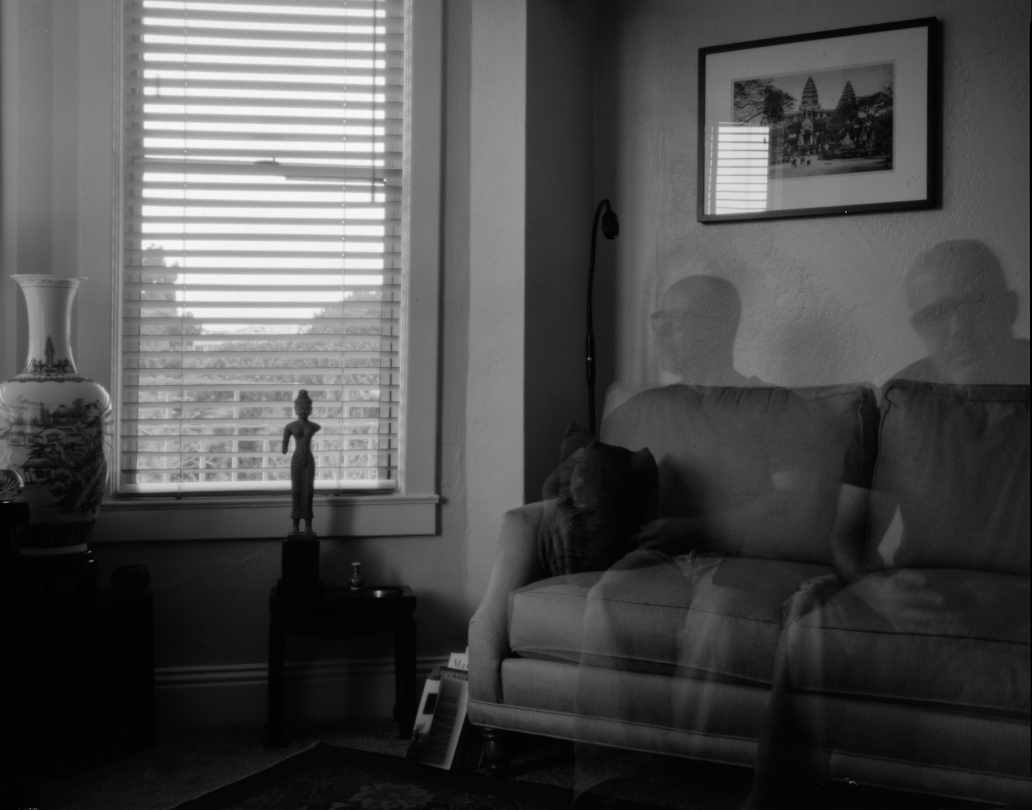 Interior space with two ghostly figures on sofa, framed artwork on wall, sculptural objects, and venetian blind light patterns