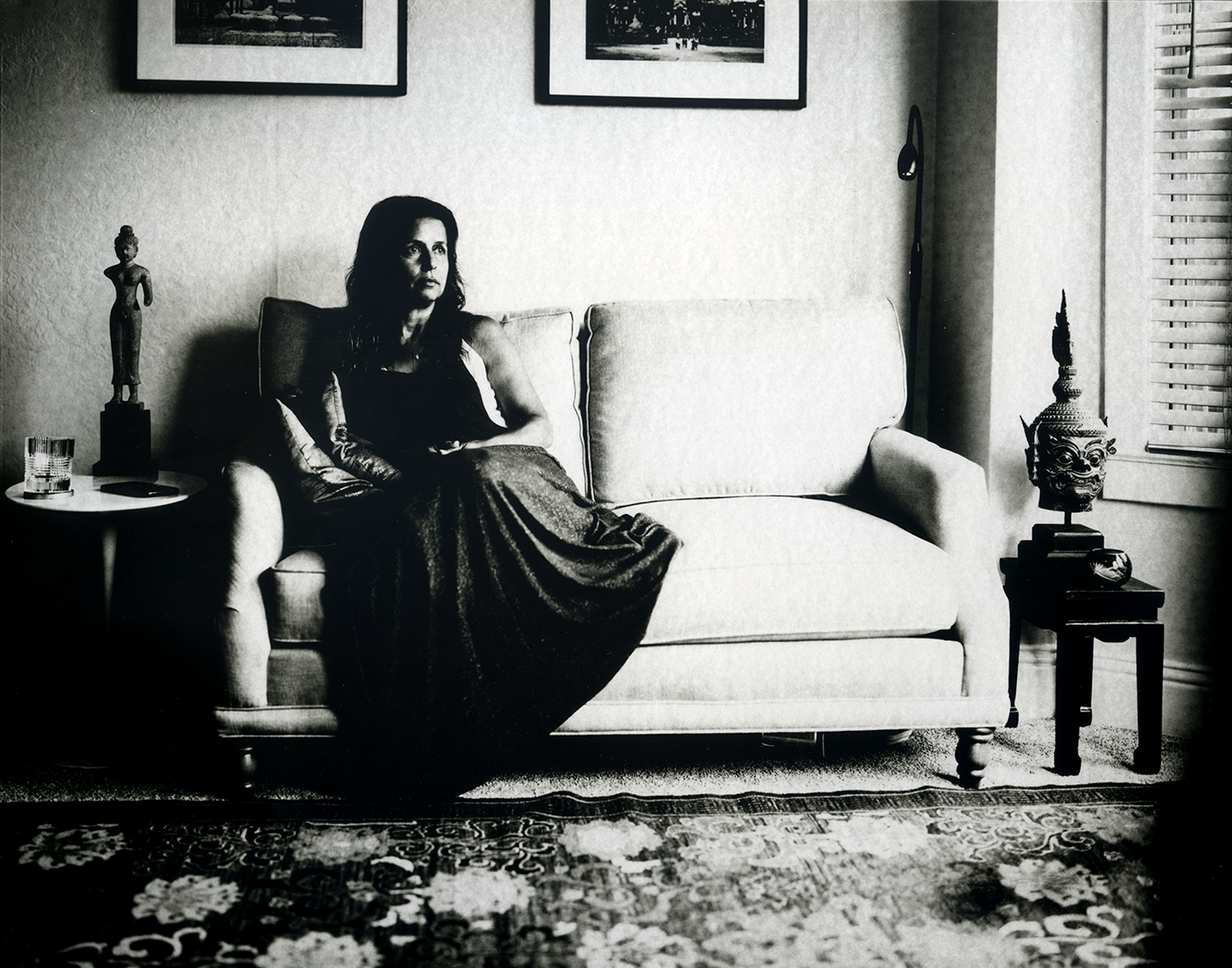 Woman in elegant black dress seated on modernist sofa in mid-century interior with framed artwork, decorative objects, and venetian blinds