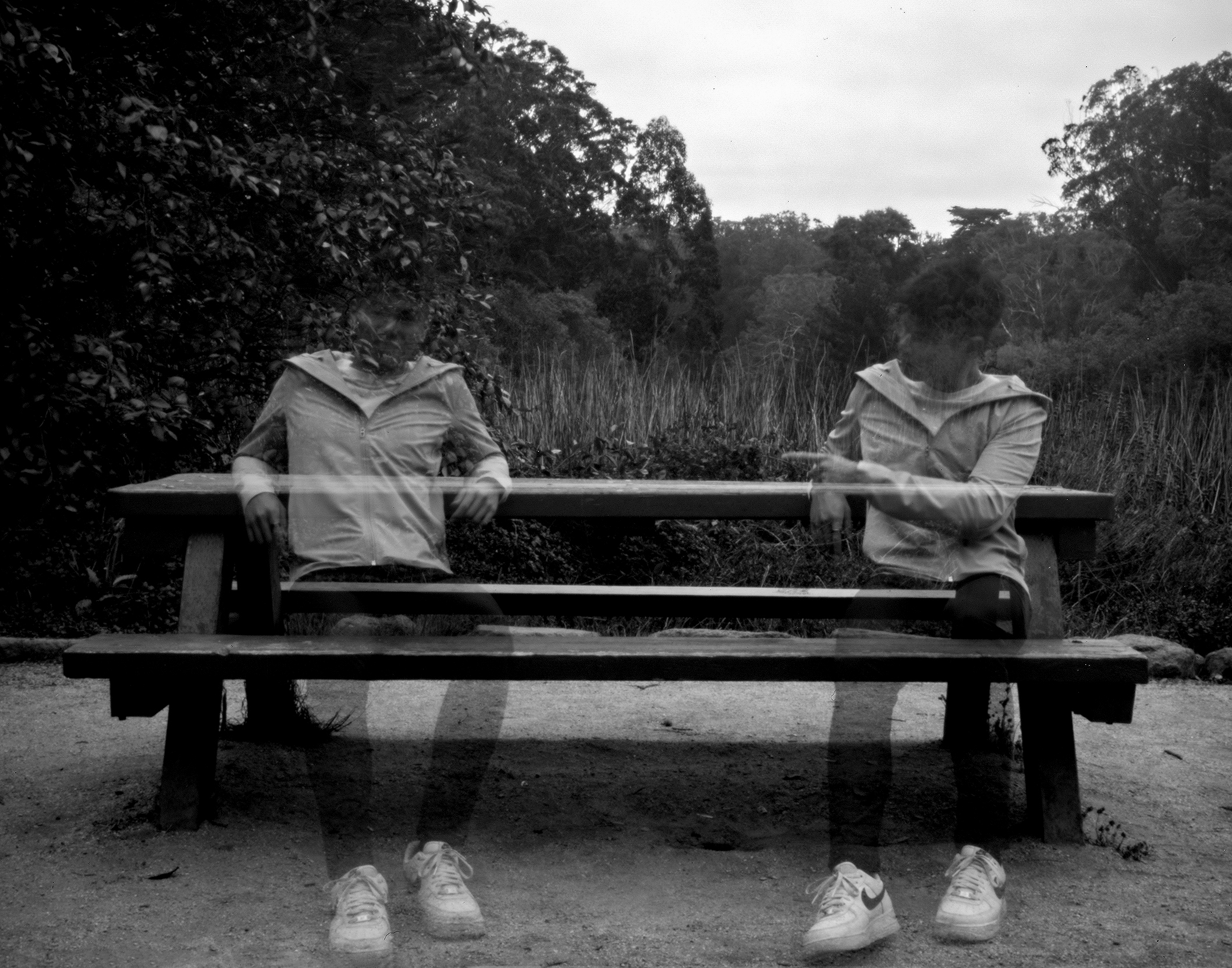 Two headless figures sitting on bench in natural landscape with shoes in foreground and dense foliage background