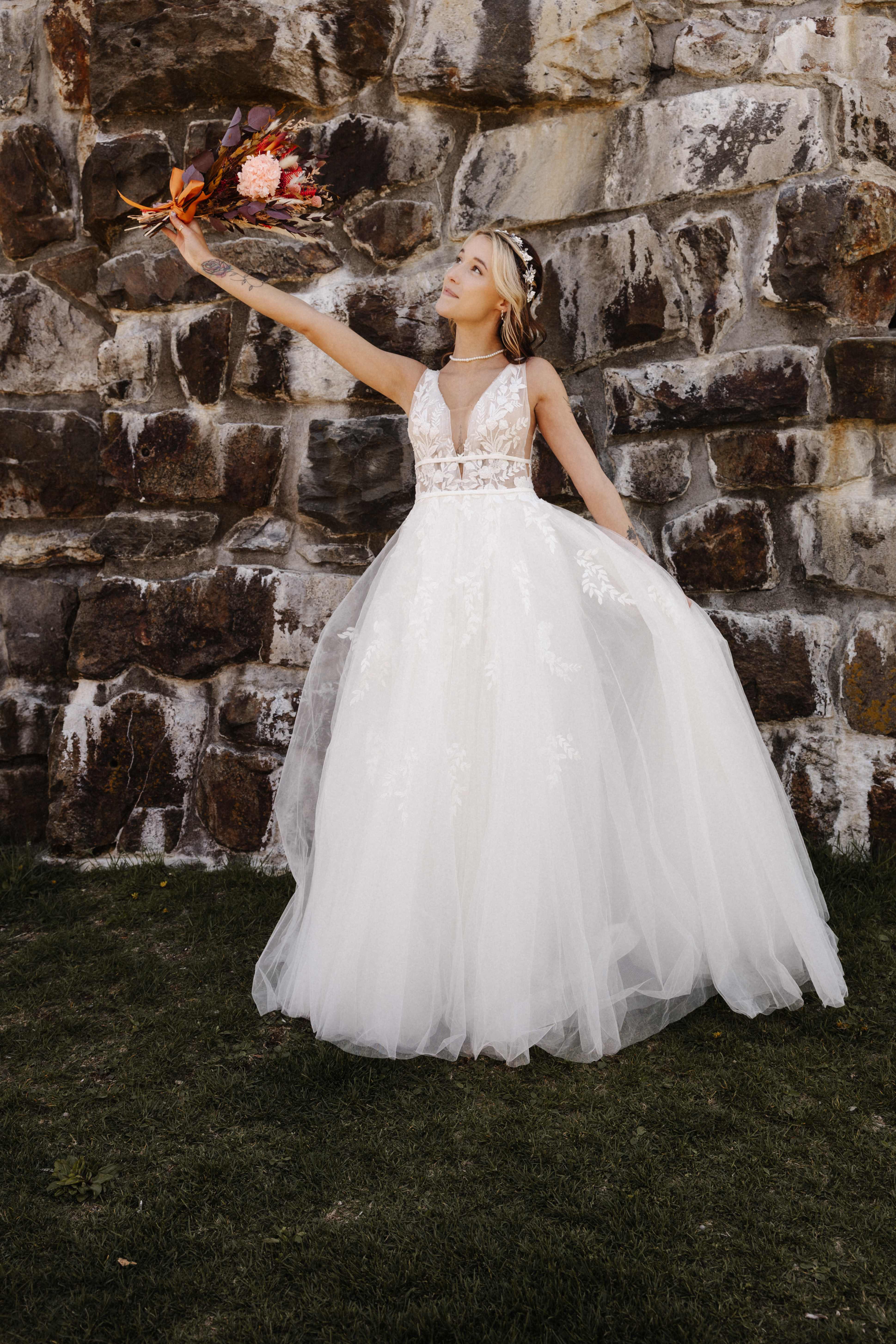 Bride raising bouquet by stone wall in ballgown dress