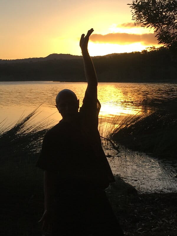 Silhouette of Kadama practising qigong by a lake at sunset, one arm raised in a flowing pose