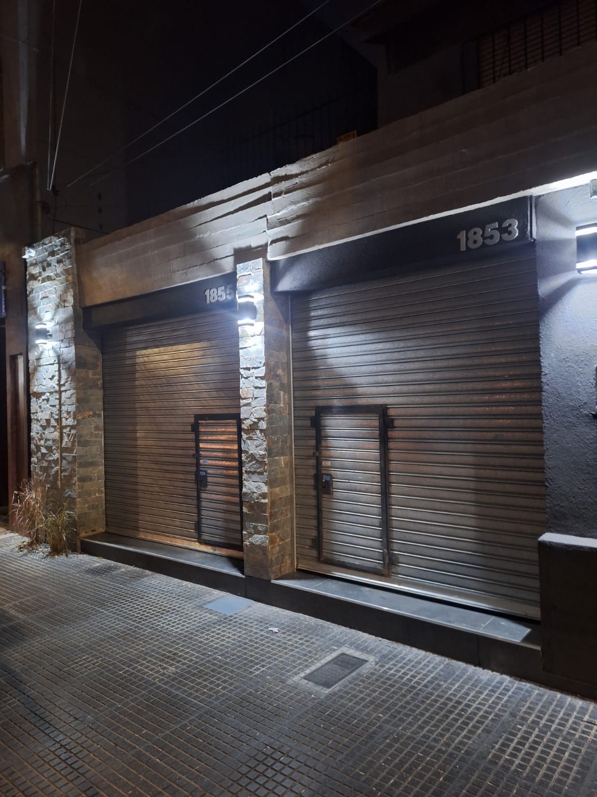 Fachada nocturna con cortinas metalicas, piedra laja iluminada y apliques de luz calida