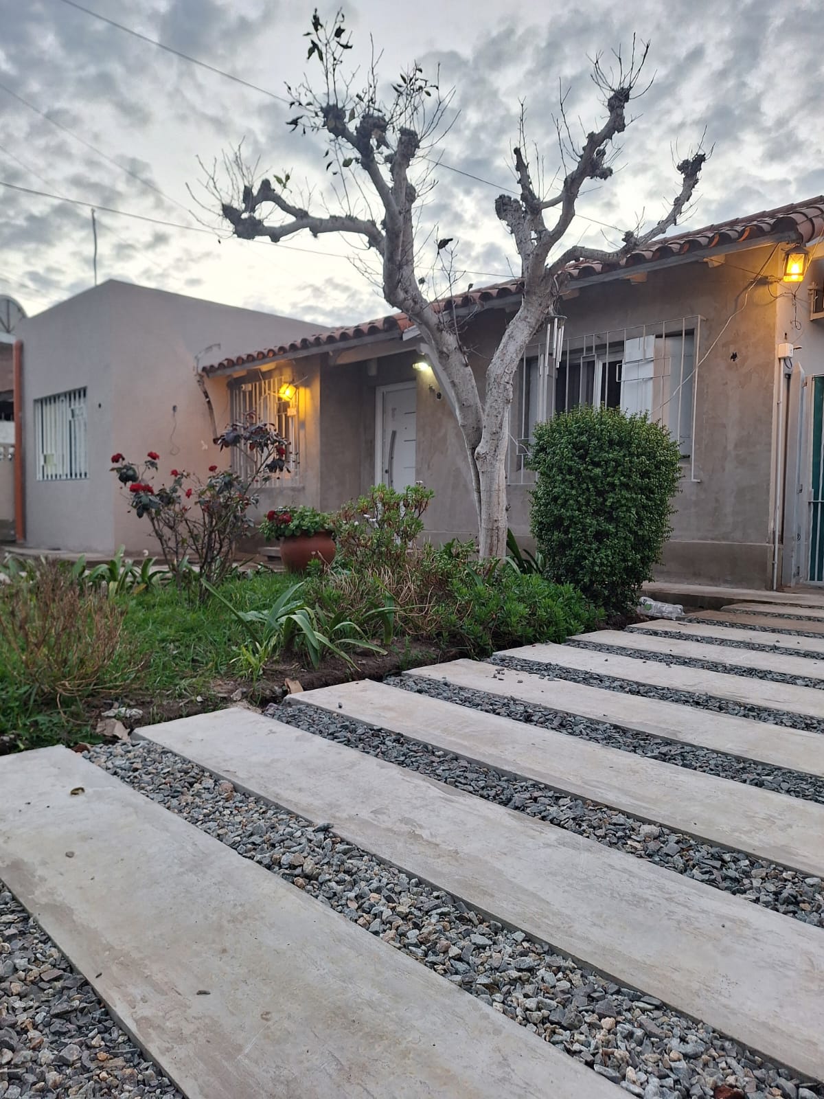 Vista exterior al atardecer con sendero de hormigón, jardín y luminarias encendidas