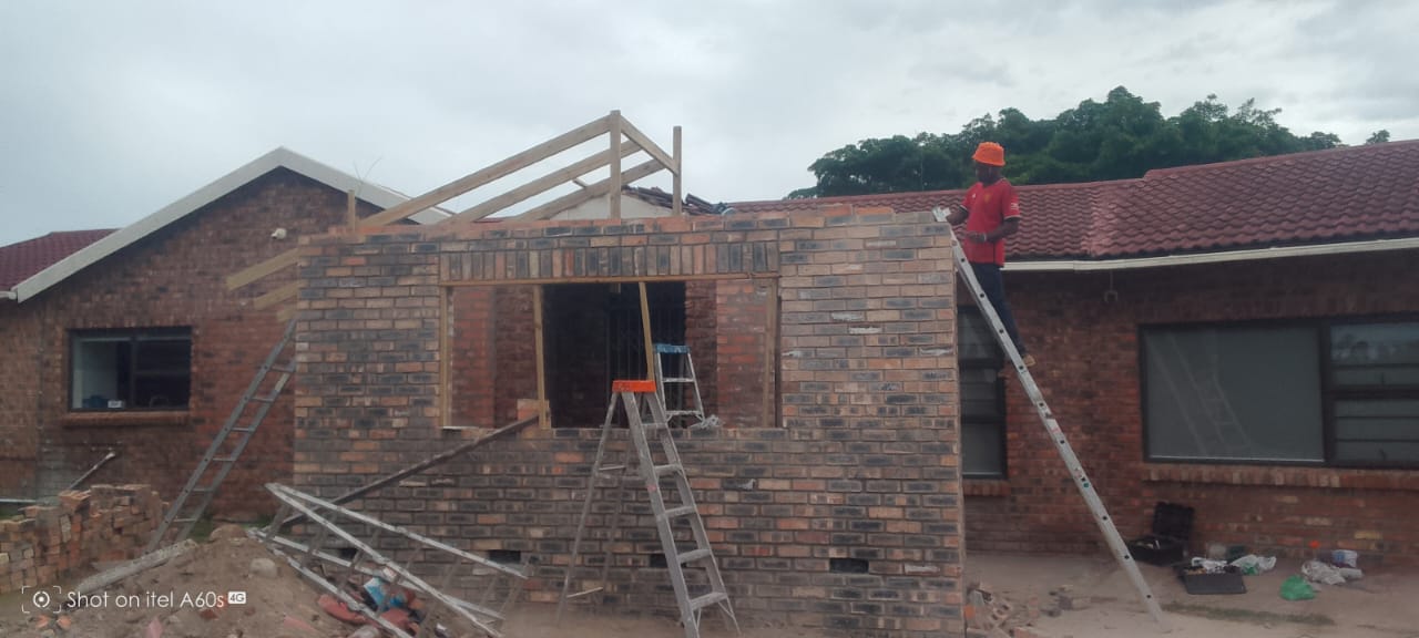 Worker in orange helmet on ladder against brick room with wooden roof trusses partially up, roof framing in progress