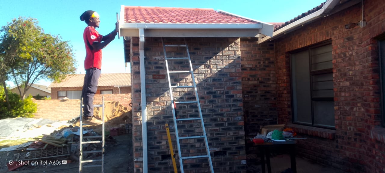 Worker in red shirt on ladder installing white PVC fascia and guttering on completed tiled roof, blue sky