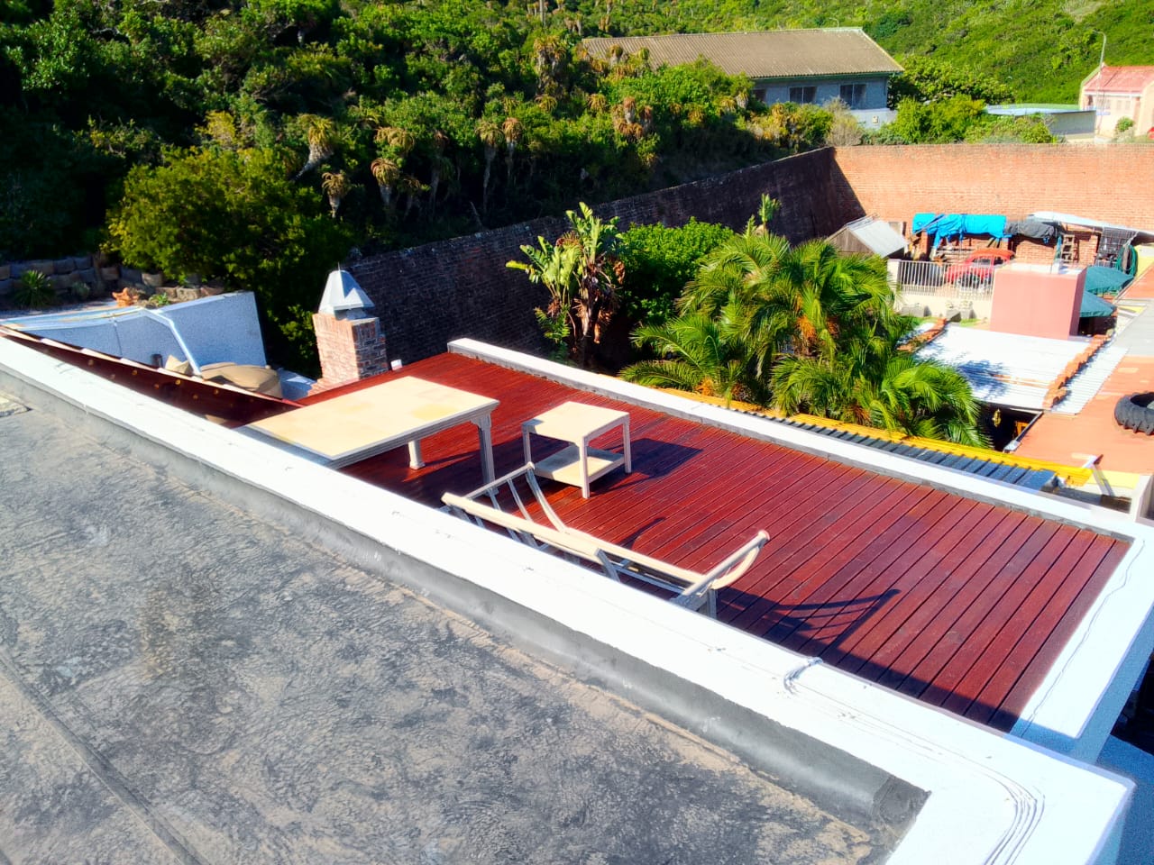 Aerial view of completed rooftop deck fully stained rich mahogany red, two white recliner chairs, palm trees and brick garden wall behind